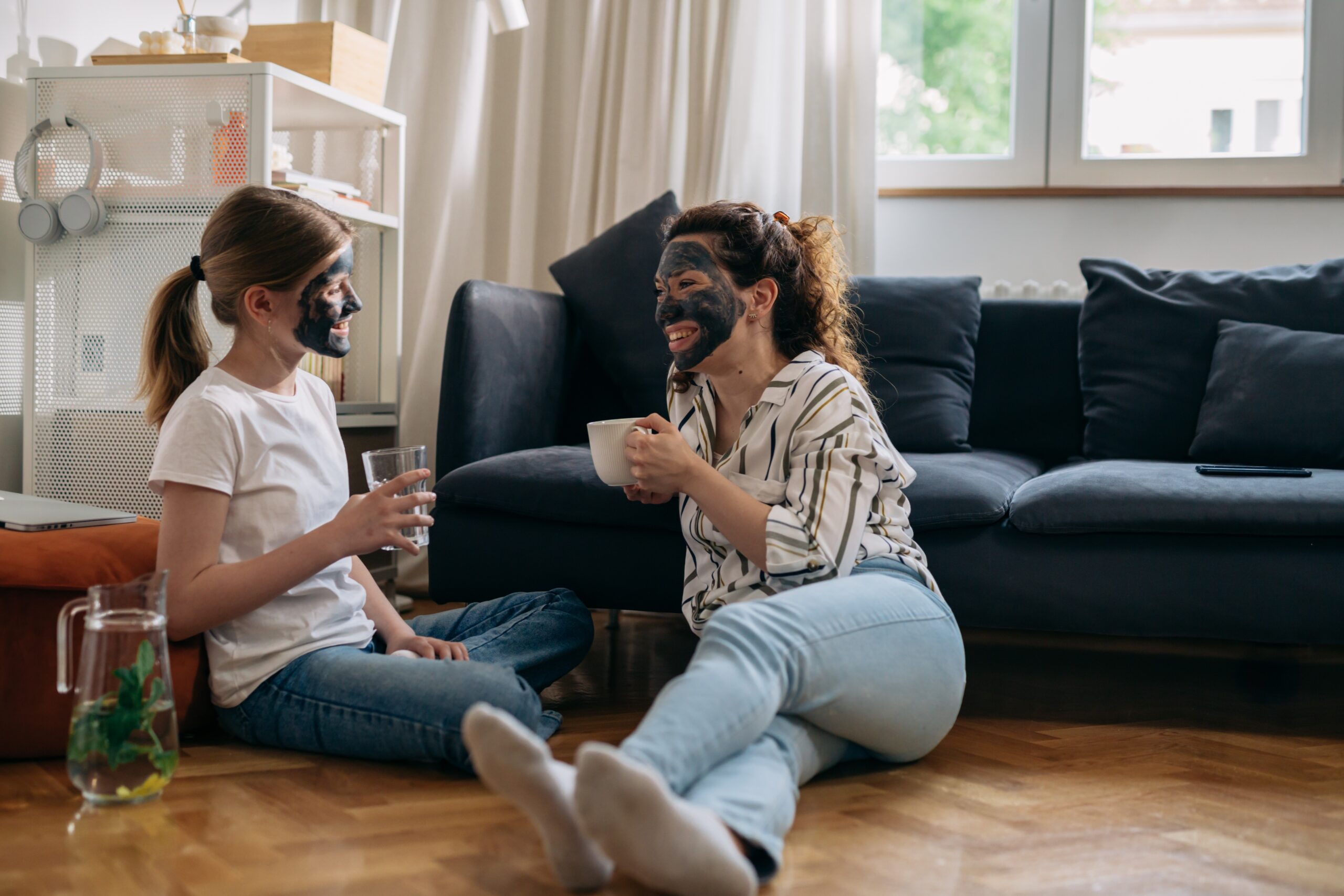 Mama a dcéra relaxujú spolu v obývačke, obe majú nanesenú prírodnú pleťovú masku. Mama drží šálku čaju, dcéra pohár vody, a obe sa usmievajú a rozprávajú.
